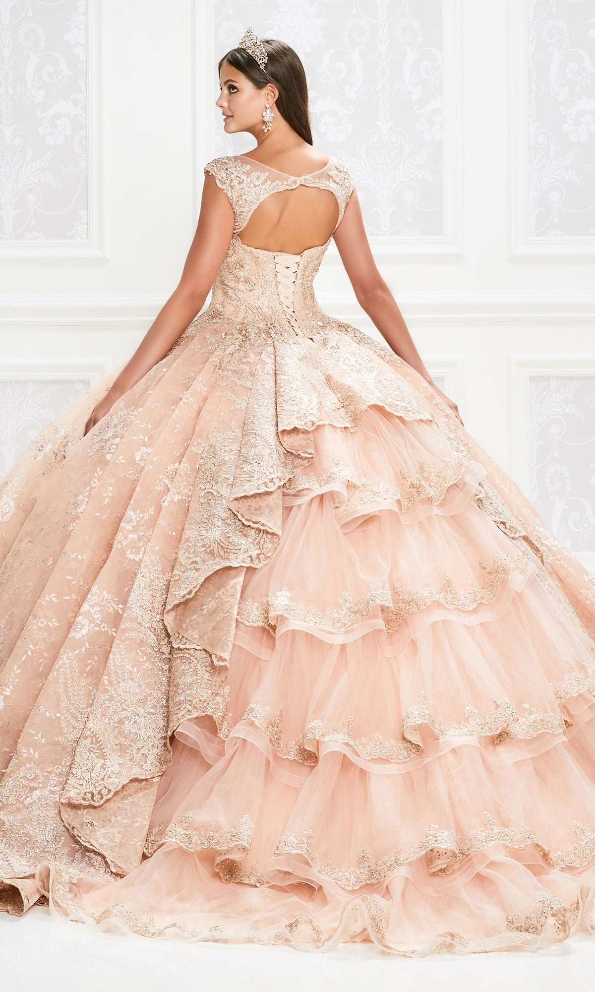 A woman in the Princesa Traditional Lace Quinceañera Dress PR12002, a light pink layered ball gown with lace and sequin accents and a corset back, wears a tiara as she stands in an ornate white room facing away.