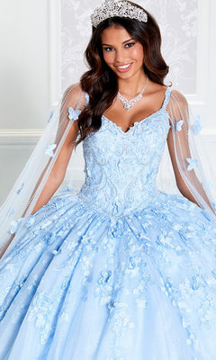 A woman wearing the Glow-in-the-Dark Princesa Quinceañera Dress PR12261 with floral details, a crystal necklace, and a tiara smiles in front of a decorative white wall.