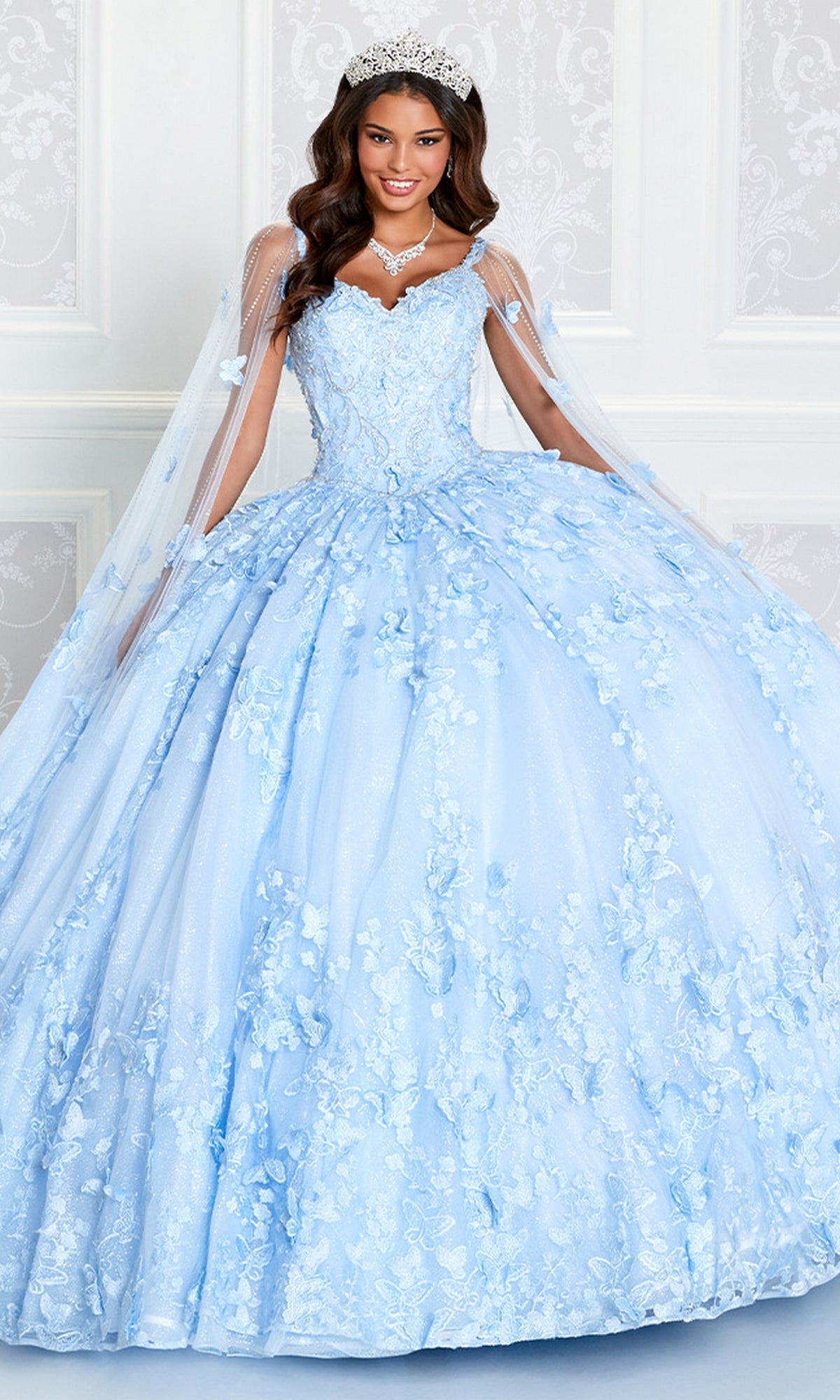 A young woman in the Glow-in-the-Dark Princesa Quinceañera Dress PR12261 by Ariana Vara, adorned with floral details, a tiara, and a necklace, smiles at the camera while standing before a white ornate wall.