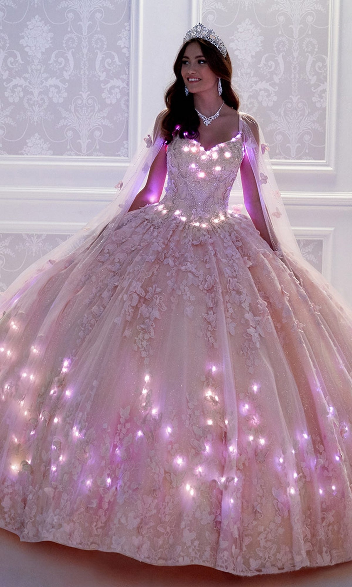 A woman in a tiara and necklace wears the Glow-in-the-Dark Princesa Quinceañera Dress PR12261 with glowing lights, standing before an ornate white wall.