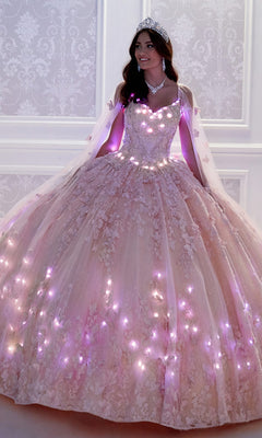 A woman in a tiara and necklace wears the Glow-in-the-Dark Princesa Quinceañera Dress PR12261 with glowing lights, standing before an ornate white wall.