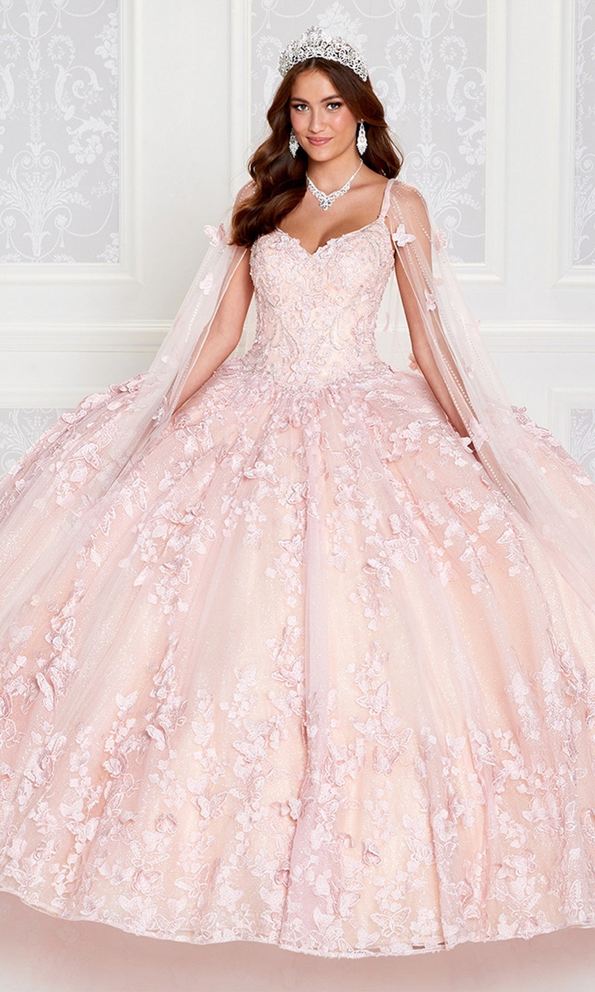 A young woman in the Glow-in-the-Dark Princesa Quinceañera Dress PR12261 by Ariana Vara, featuring pink floral details and off-shoulder tulle sleeves, stands with a tiara and jewelry before a white ornate wall.