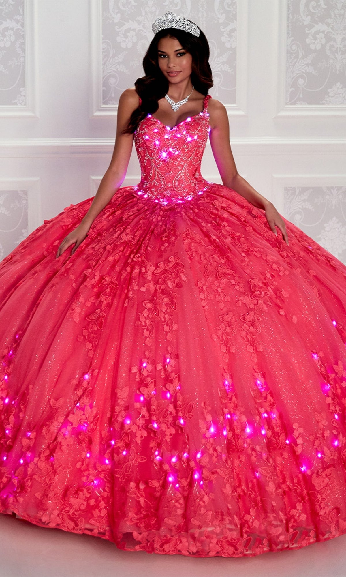 A woman in a sparkling, bright pink Glow-in-the-Dark Princesa Quinceañera Dress PR12261 with LED lights, wearing a tiara and necklace, stands against a white ornate wall.