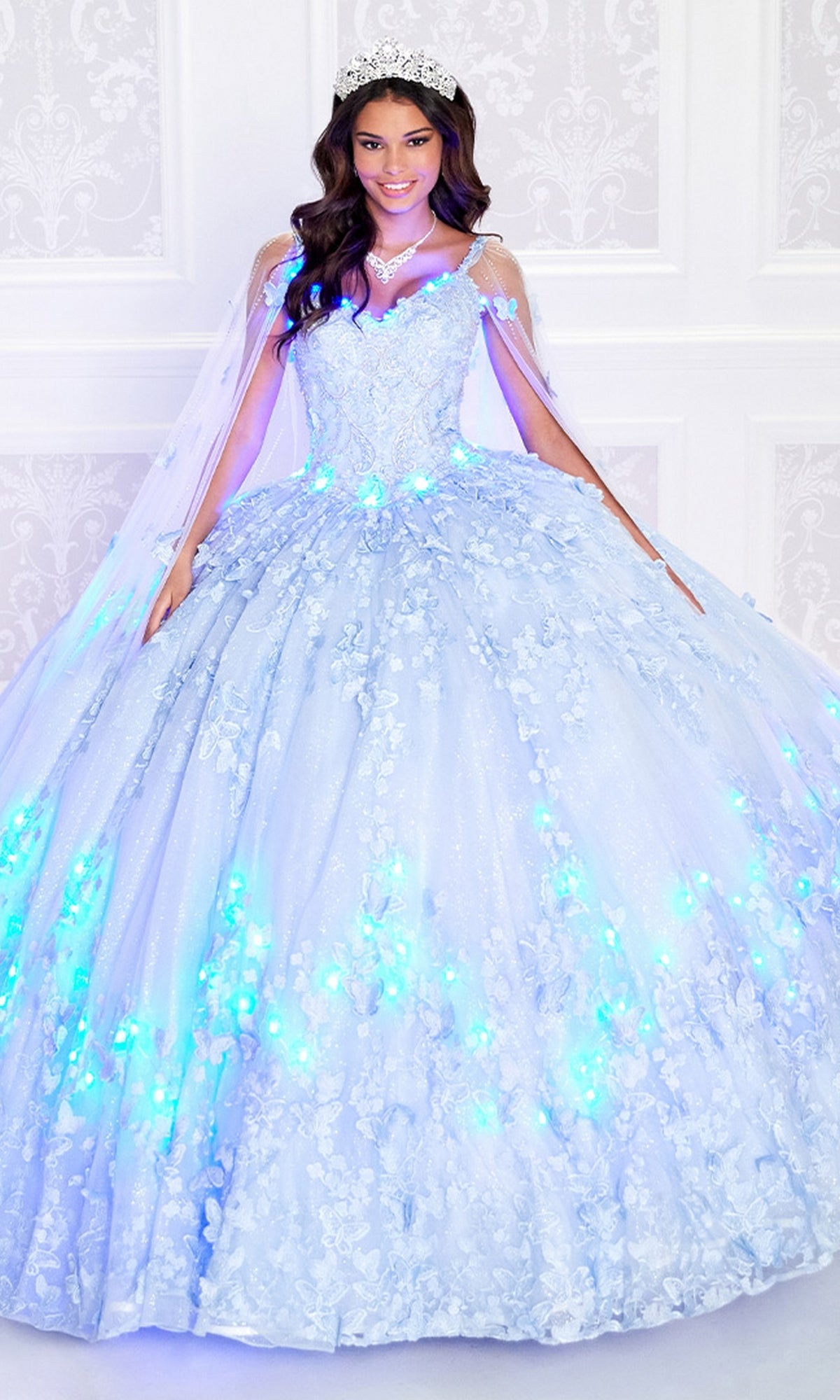 A woman in the Glow-in-the-Dark Princesa Quinceañera Dress PR12261, featuring a glowing light blue ball gown with floral details, tiara, and necklace, stands before a white background. This radiant look is inspired by Princesa by Ariana Vara.