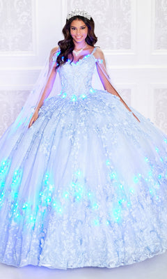 A woman in the Glow-in-the-Dark Princesa Quinceañera Dress PR12261, featuring a glowing light blue ball gown with floral details, tiara, and necklace, stands before a white background. This radiant look is inspired by Princesa by Ariana Vara.