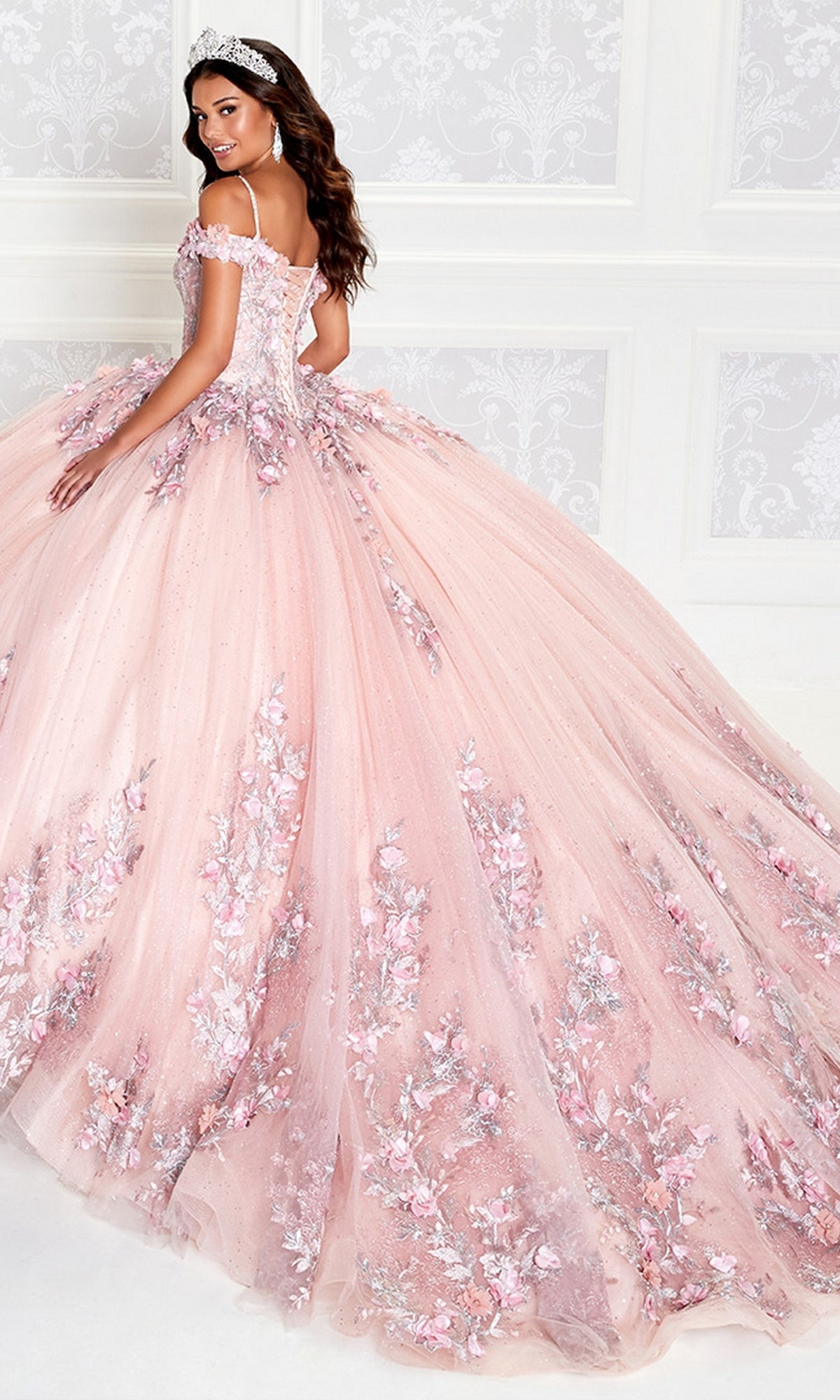 A woman wears the Princesa Quinceañera Dress PR12263, featuring off-the-shoulder styling and 3-D floral embellishments, paired with a tiara as she stands gracefully before a white ornate wall.