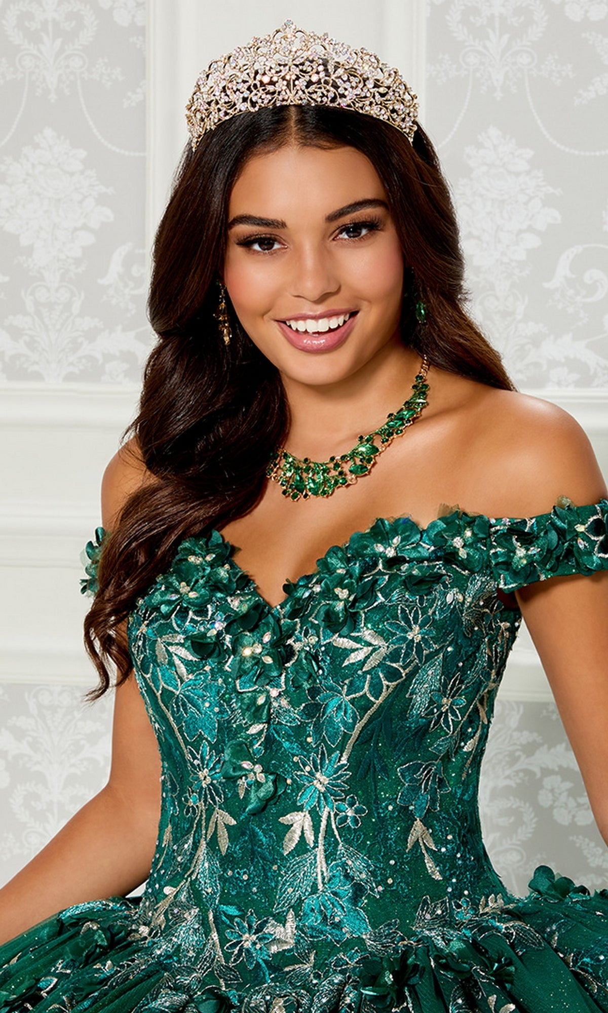 A young woman smiles in front of a light decorative background, wearing the Princesa Quinceañera Dress PR12263 with 3-D flowers, a jeweled tiara, and a matching green necklace.