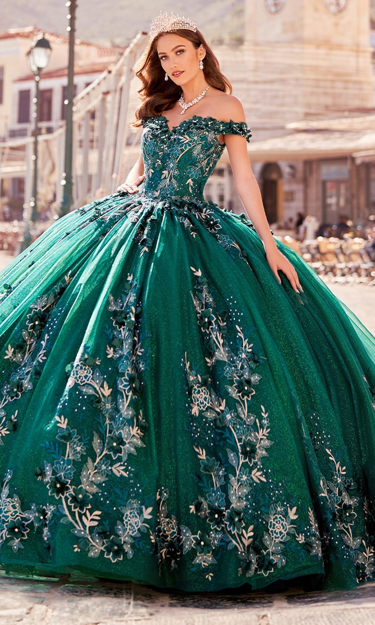 A woman wearing the Princesa Quinceañera Dress PR12263 with 3-D Flowers, in emerald green lace with silver floral details, a tiara, and necklace, stands outdoors before historic buildings.