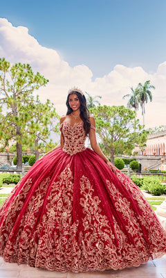 A young woman wearing a crown and the PR12264 Princesa Quince Dress with a glitter tulle skirt stands outdoors in a formal garden, with trees and classical architecture in the background.