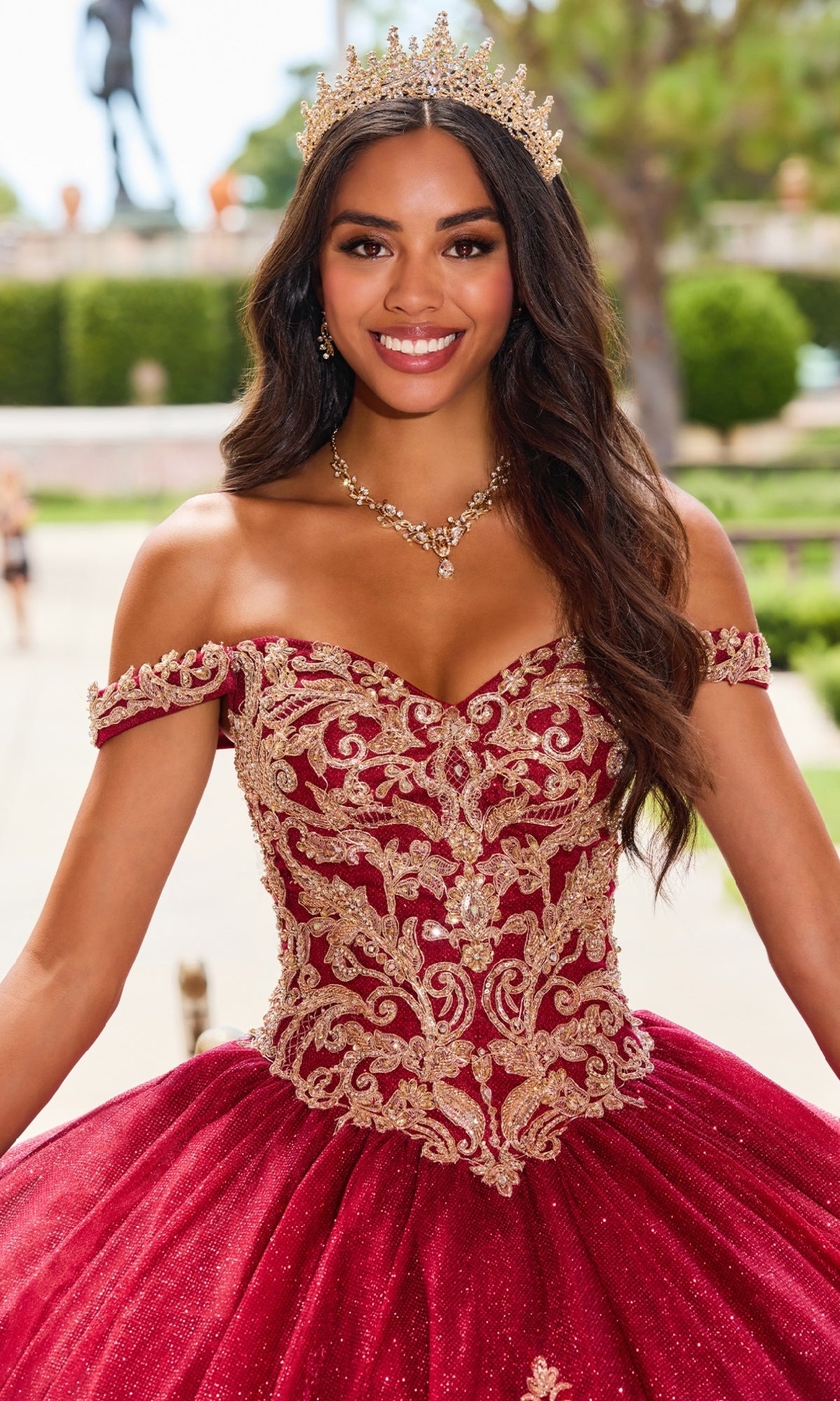 A woman in the PR12264 Princesa Quince Dress with Glitter Tulle wears a gold crown and stands outdoors, smiling at the camera in her red, off-the-shoulder, embroidered gown.