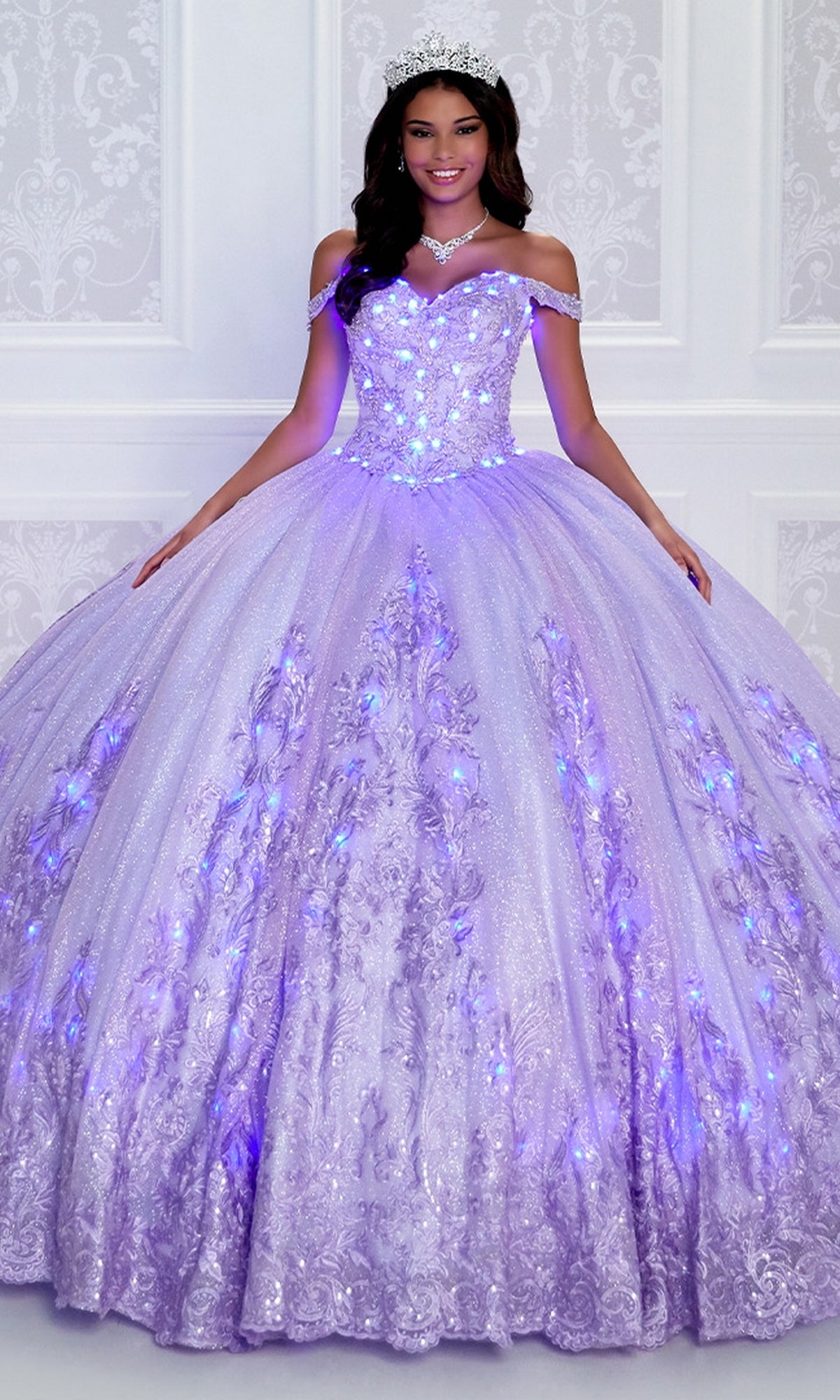 A young woman in the PR12264 Princesa Quince Dress with glitter tulle and glowing LED lights, topped with a tiara, stands before a white patterned wall.