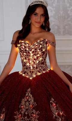 Wearing the PR12264 Princesa Quince Dress with Glitter Tulle—an ornate, off-the-shoulder maroon gown with gold embroidery, built-in lights, and a tiara—a woman stands indoors against a patterned wall.
