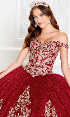 A young woman poses indoors against a light patterned wall, wearing the PR12264 Princesa Quince Dress with Glitter Tulle in sparkling red and gold, featuring intricate embroidery, an off-the-shoulder design, tiara, and drop earrings.