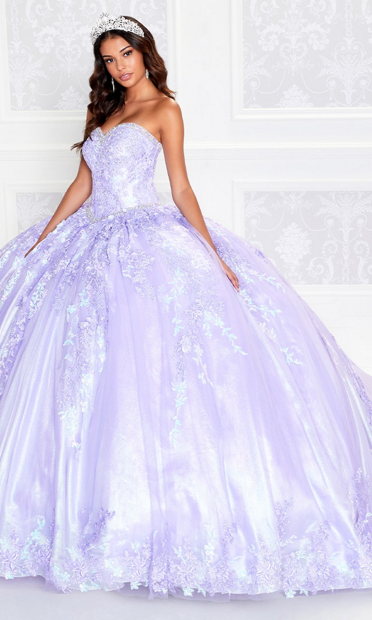 A woman wears the Strapless Princesa PR12265 Quince Dress with Train, featuring lavender floral embroidery and a tiara, standing before a white ornate wall.