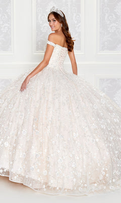 A woman in a sparkling Princesa Glow-in-the-Dark Quince Dress PR12266 with an off-the-shoulder neckline, full skirt, and tiara stands before an ornate white wall.