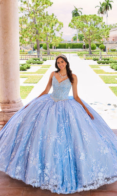 A young woman stands outdoors on a stone walkway surrounded by greenery and columns, wearing the Princesa by Ariana Vara Lace Quince Dress PR12268 in light blue.