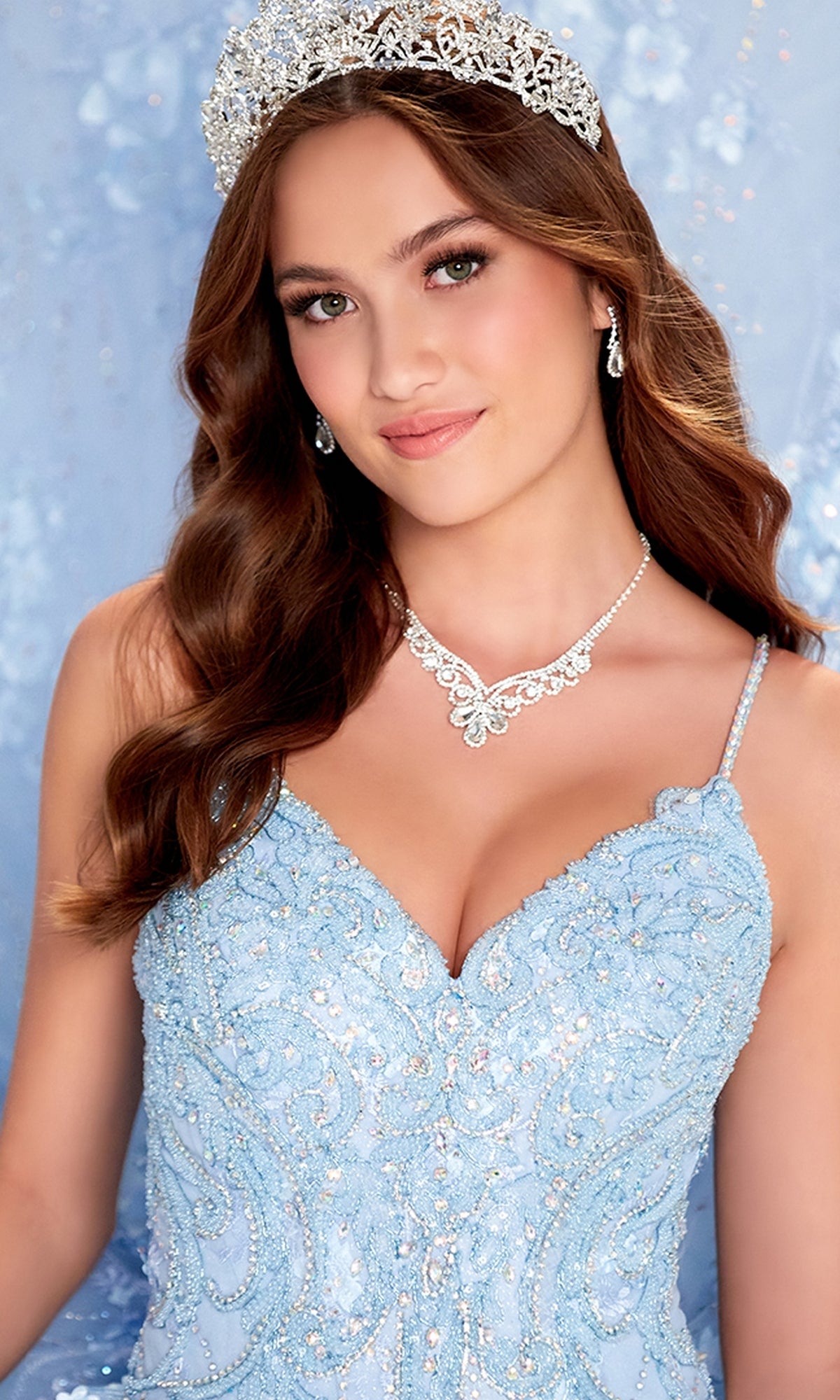 A woman poses in front of a light blue background, wearing a jeweled tiara, sparkling necklace, earrings, and the Princesa by Ariana Vara Lace Quince Dress PR12268.