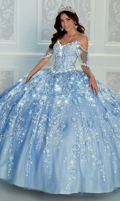 A young woman wearing the Cold-Shoulder Princesa Quinceanera Dress PR12271, featuring light blue fabric and floral embroidery, stands with a tiara in front of a pale, patterned wall.