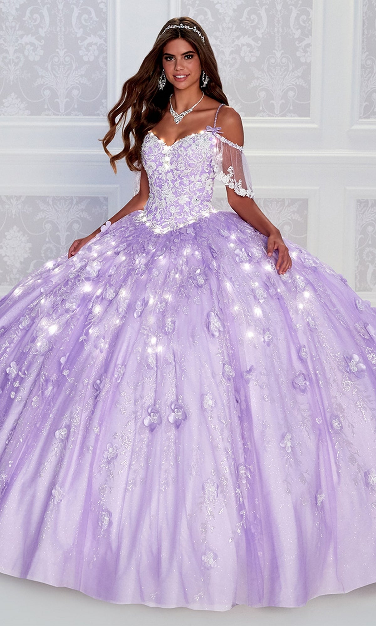 A woman wearing the Cold-Shoulder Princesa Quinceanera Dress PR12271 with sparkling lavender fabric and floral details smiles at the camera in front of a white patterned wall.