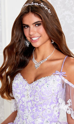 Young woman with long brown hair smiles at the camera, wearing the Cold-Shoulder Princesa Quinceañera Dress PR12271, paired with a jeweled necklace, matching earrings, and a decorative headband.