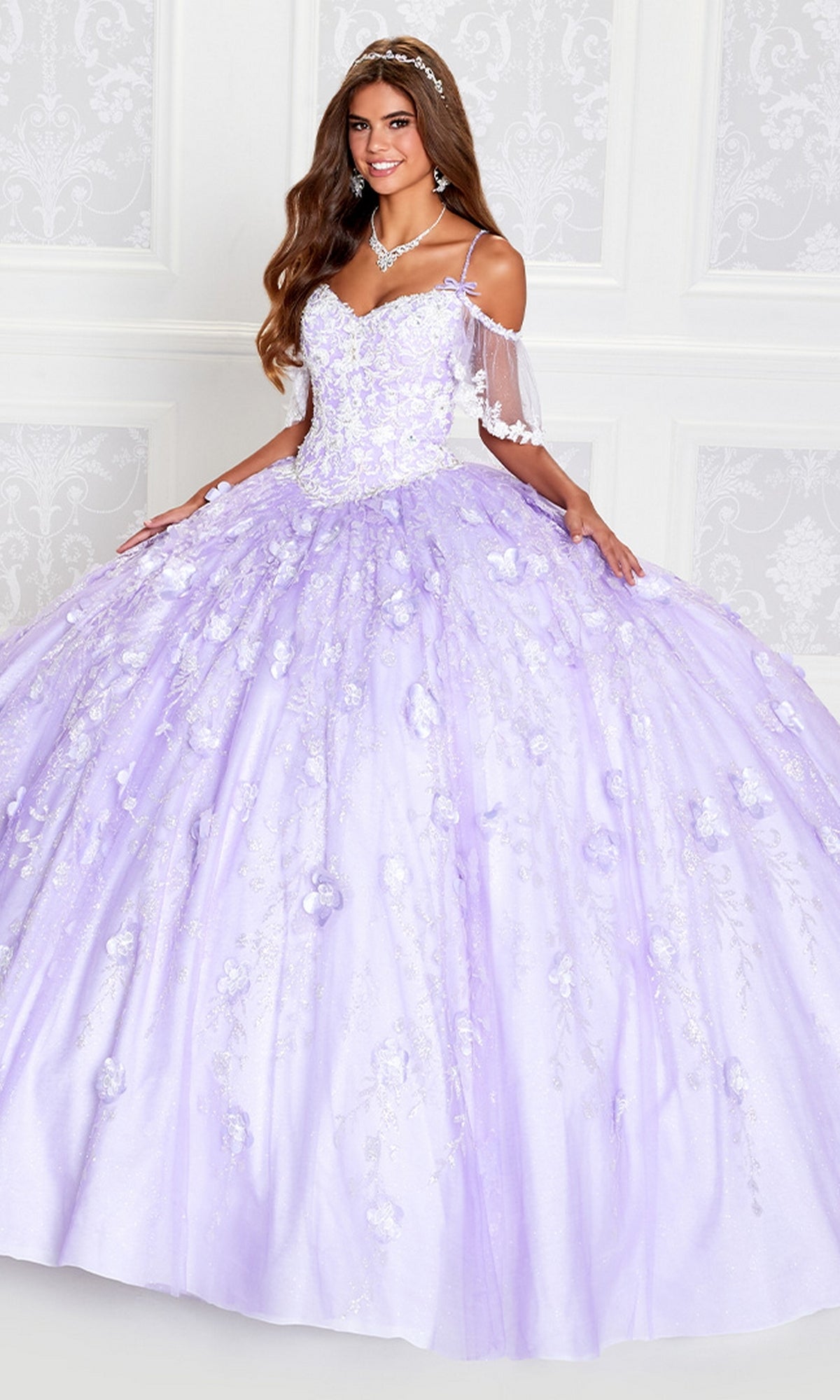 A young woman smiles indoors wearing the Cold-Shoulder Princesa Quinceanera Dress PR12271, a lilac ball gown with floral embellishments and off-the-shoulder sleeves.