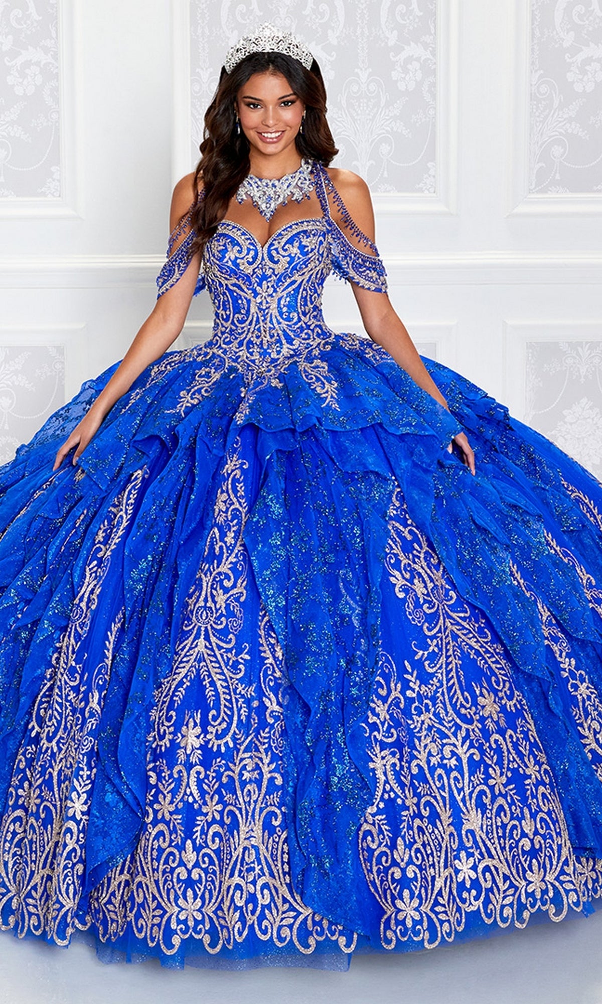 A woman in the Princesa PR12274 Quinceañera Dress with Glitter and gold embroidery, accessorized with a tiara, stands before a white wall, showcasing the dazzling elegance of this Ariana Vara design.