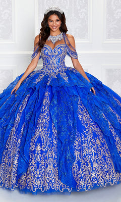A woman in the Princesa PR12274 Quinceañera Dress with Glitter and gold embroidery, accessorized with a tiara, stands before a white wall, showcasing the dazzling elegance of this Ariana Vara design.
