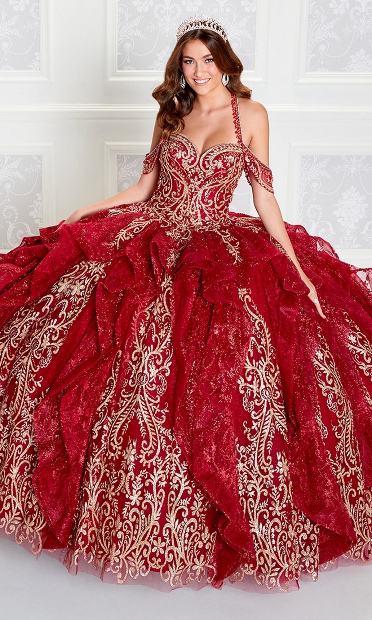 A woman in a tiara poses in the elegant Princesa PR12274 Quinceañera Dress with glitter, featuring gold embroidery and off-the-shoulder straps, set against an ornate white wall.