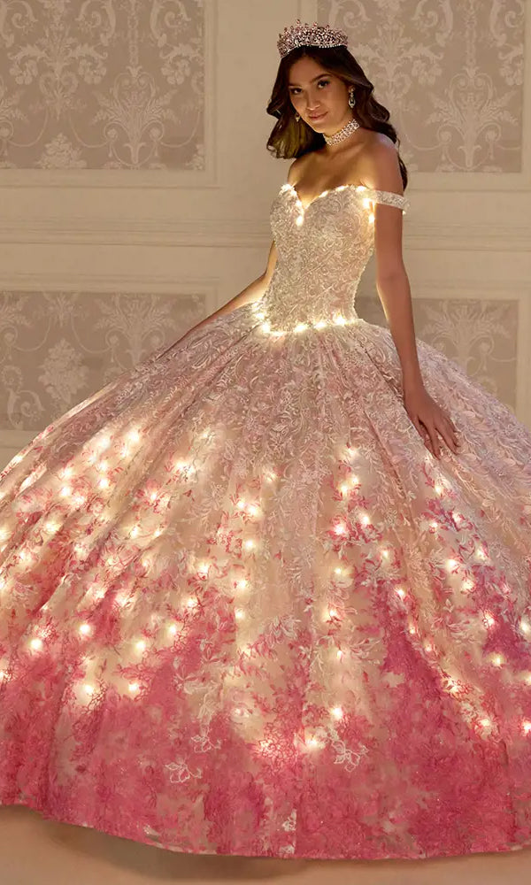 A woman poses indoors in the PR12275 Princesa Quinceañera Dress, a pink and white embroidered lace gown with built-in lights, accessorized with a tiara and choker necklace.