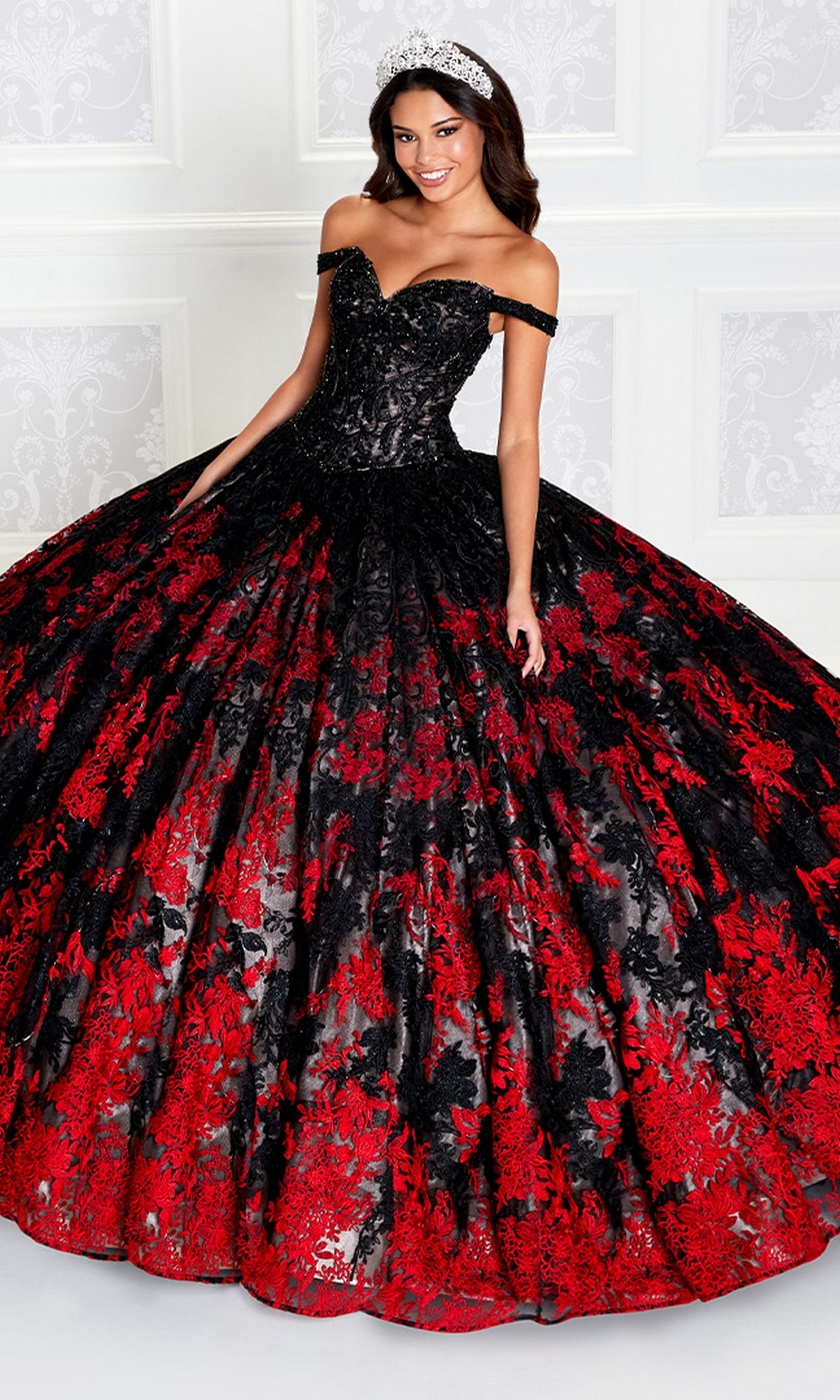 A woman wears the PR12275 Princesa Quinceañera Dress featuring an off-the-shoulder embroidered lace bodice and a voluminous black and red floral skirt, standing against a white paneled background.