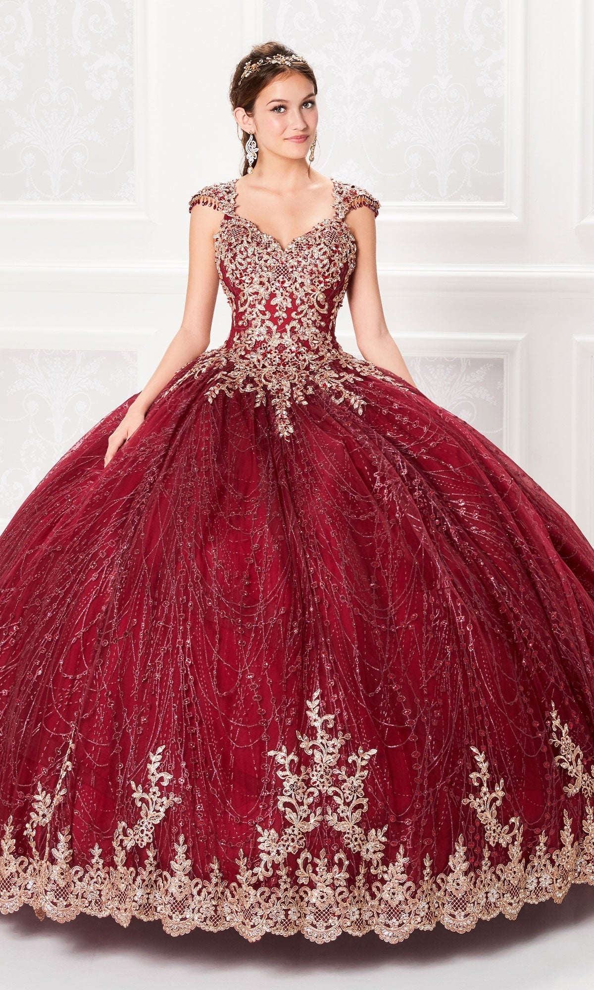 A young woman in the Cap-Sleeve Quinceanera Dress: Princesa PR21964, featuring deep red fabric, gold embroidery, and an embellished corset bodice, stands indoors against a white ornate wall.
