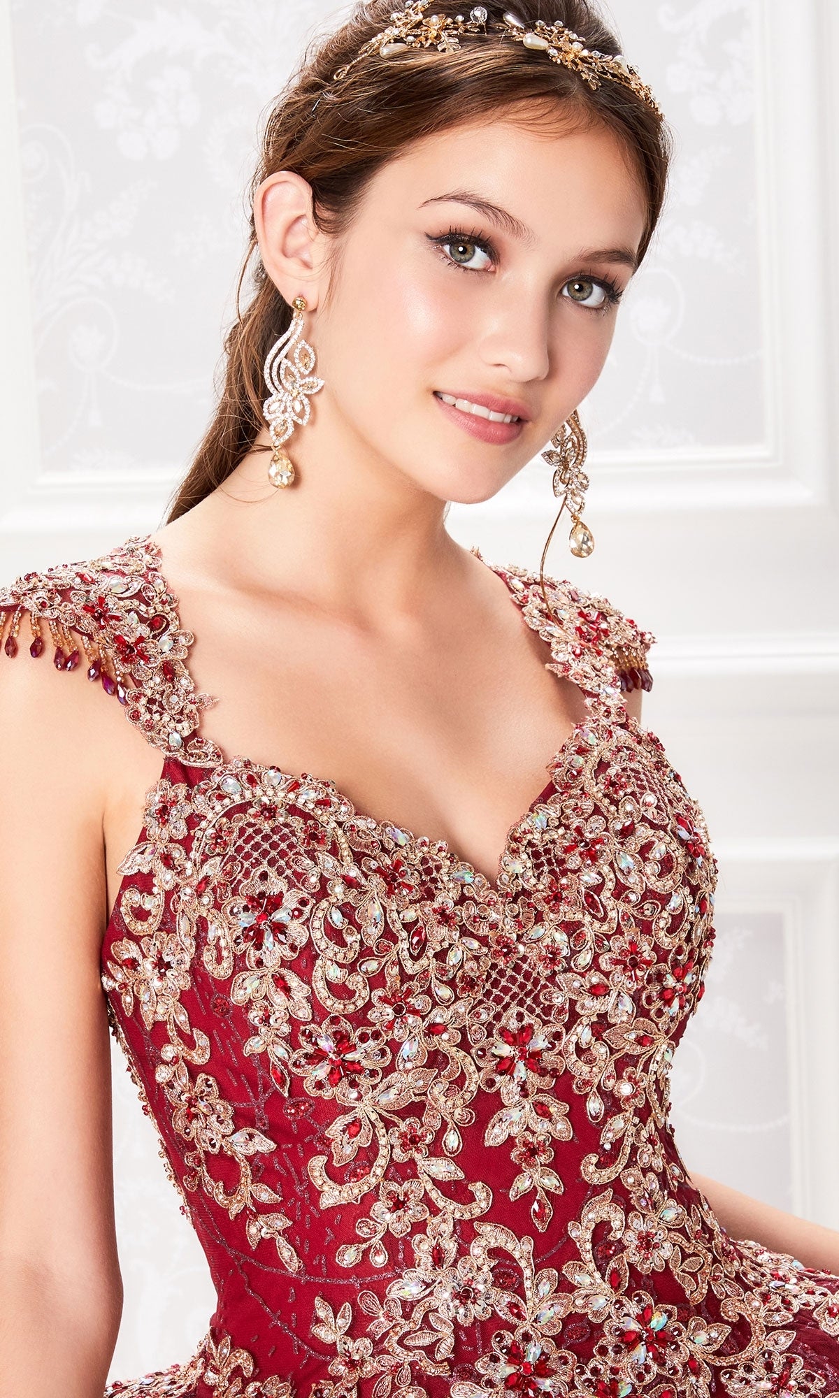 A young woman poses in front of a white patterned background wearing the Princesa PR21964 Cap-Sleeve Quinceanera Dress with a corset bodice, matching earrings, and a tiara.