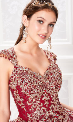 A young woman poses in front of a white patterned background wearing the Princesa PR21964 Cap-Sleeve Quinceanera Dress with a corset bodice, matching earrings, and a tiara.