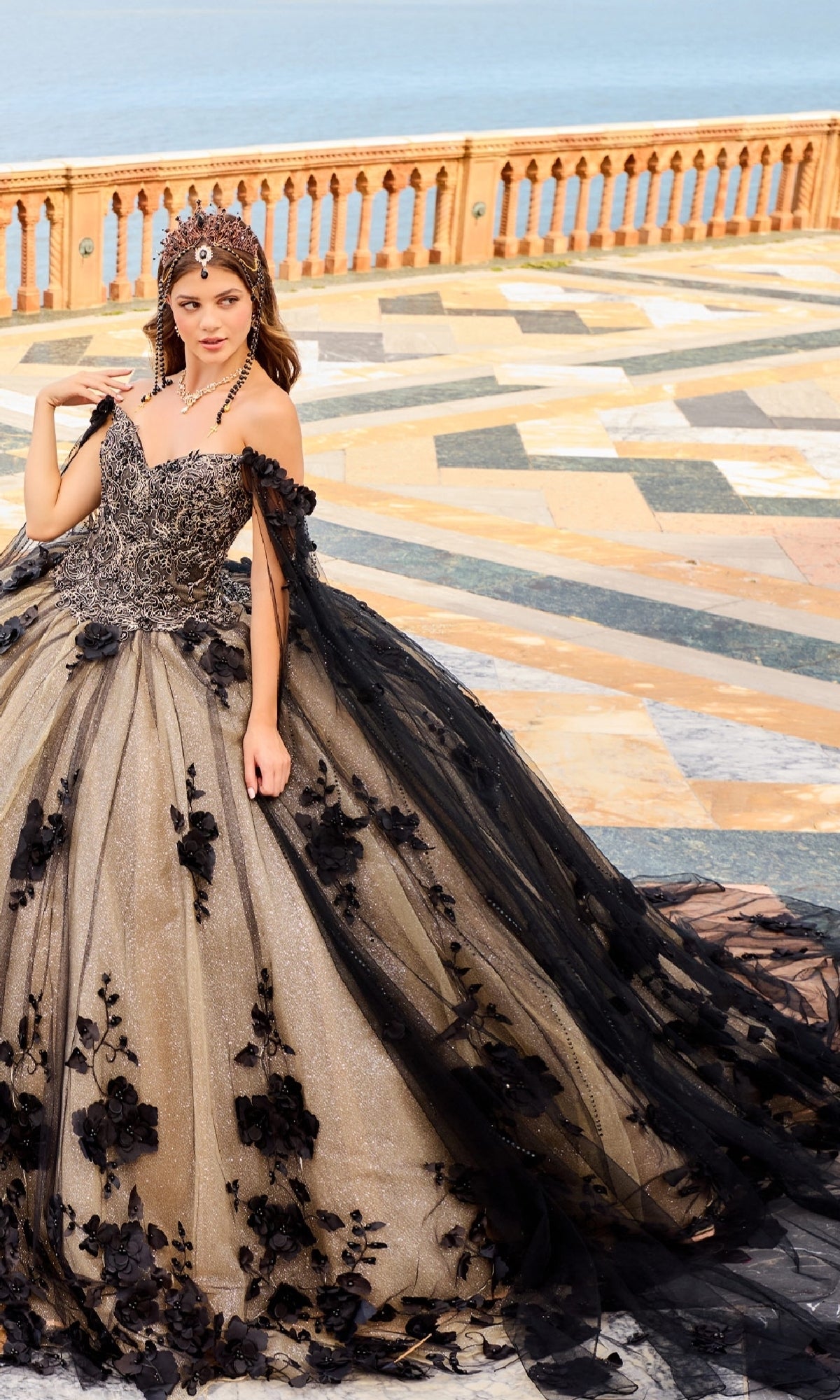 A woman stands outdoors in the PR22021 Princesa Glow-in-the-Dark Quinceañera Dress, featuring black and gold details, floral appliqués, and a matching headpiece. Behind her is a patterned stone terrace with a sea view.