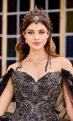 A woman poses gracefully by a grid window in the PR22021 Princesa Glow-in-the-Dark Quinceañera Dress, an ornate black and gold gown adorned with intricate embroidery, a jeweled crown, and a matching necklace.