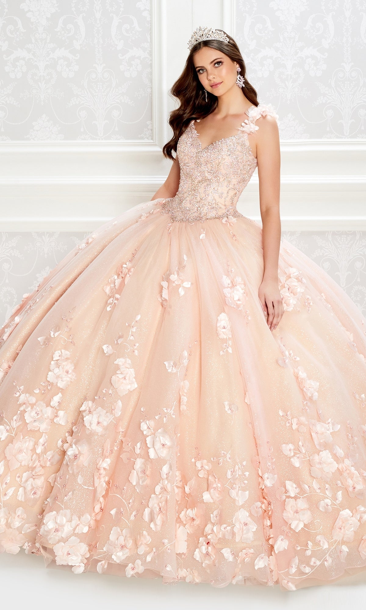 A woman wearing the PR22021 Princesa Glow-in-the-Dark Quinceañera Dress and a tiara stands in front of an ornate white wall.
