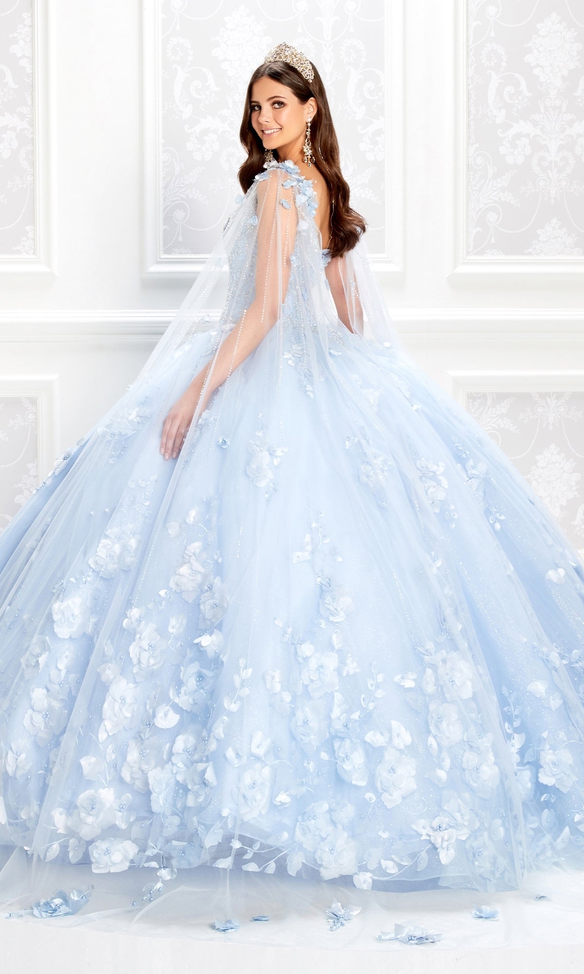 A woman in a light blue PR22021 Princesa Glow-in-the-Dark Quinceañera Dress with 3D floral accents and a sheer cape poses in an ornate white room.