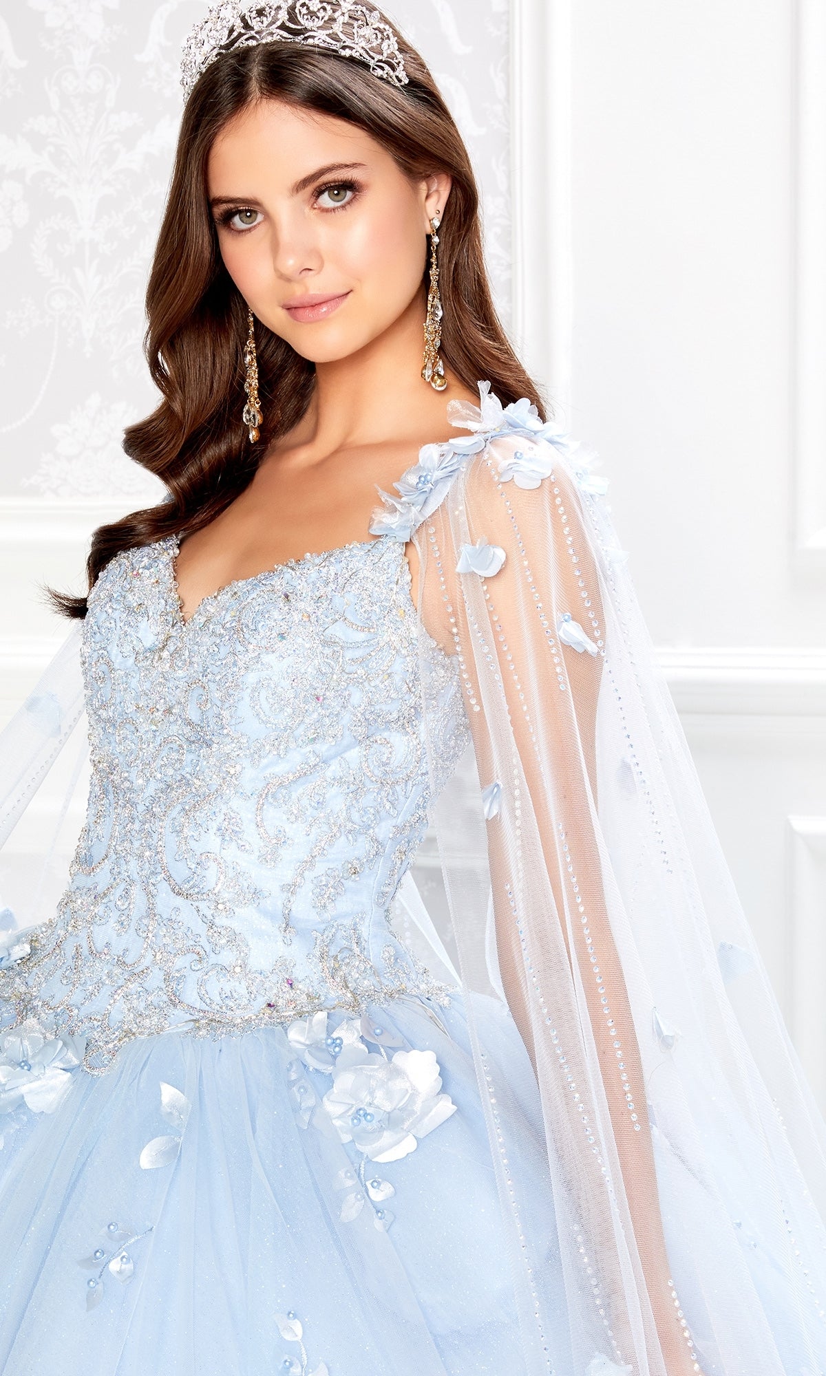 A young woman wearing the PR22021 Princesa Glow-in-the-Dark Quinceañera Dress with sheer sleeves and floral details stands indoors, facing the camera.