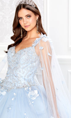 A young woman wearing the PR22021 Princesa Glow-in-the-Dark Quinceañera Dress with sheer sleeves and floral details stands indoors, facing the camera.