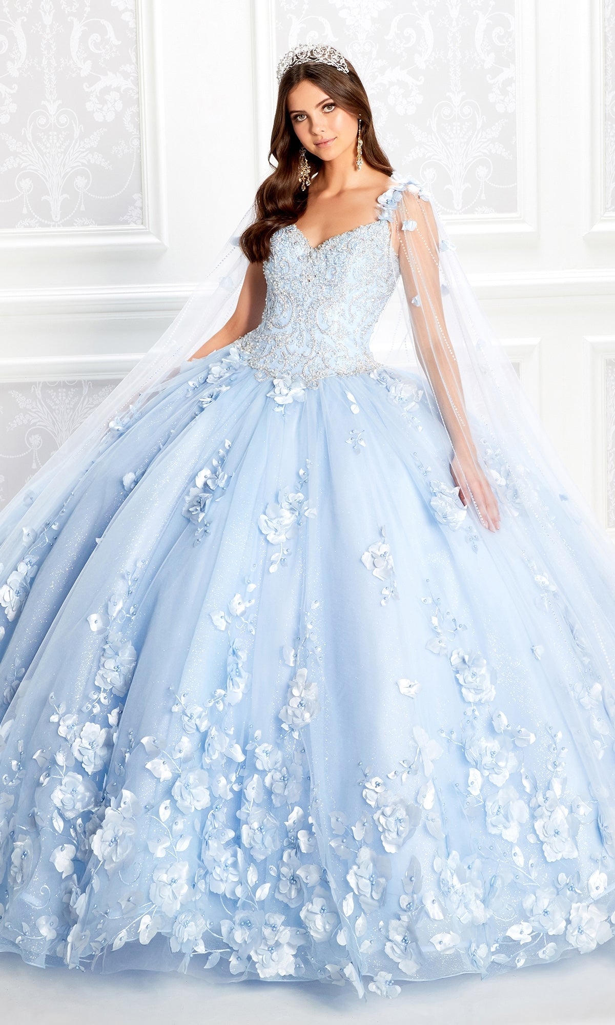 A woman in the PR22021 Princesa Glow-in-the-Dark Quinceañera Dress with floral appliqués stands in a decorated white room, wearing a tiara and radiating elegance in this Ariana Vara-inspired look.