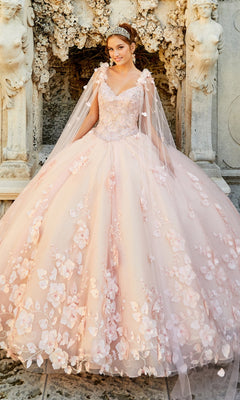 A young woman in the PR22021 Princesa Glow-in-the-Dark Quinceañera Dress with floral details and sheer shoulder drapes stands before an ornate stone structure, wearing a tiara and smiling.