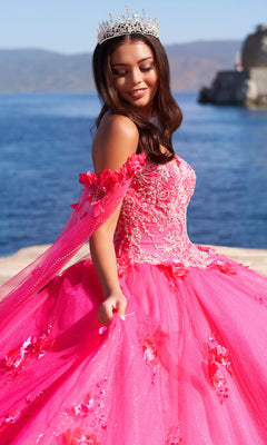 A woman in the PR22021 Princesa Glow-in-the-Dark Quinceañera Dress and a tiara stands by a waterfront, with blue water and distant hills in the background.