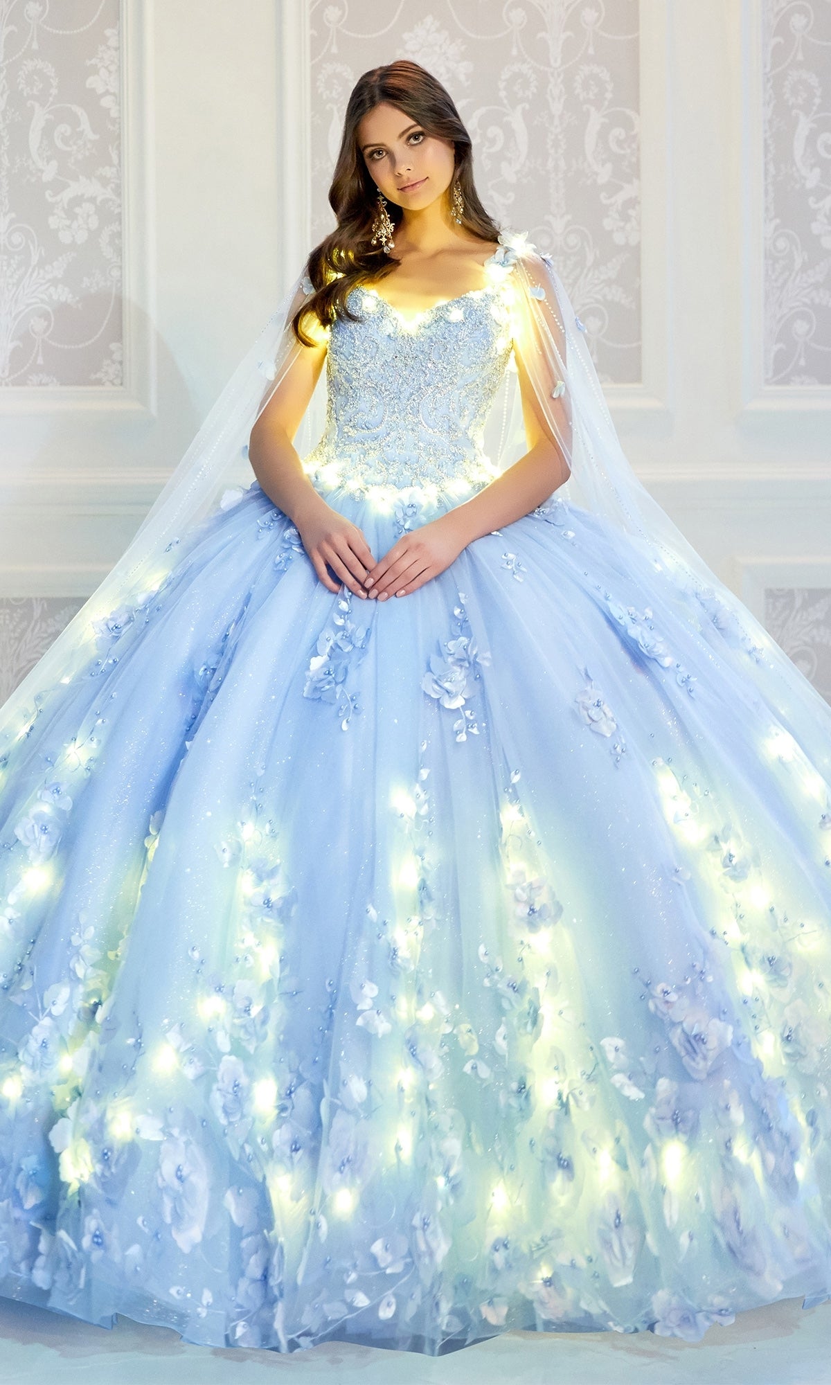 A young woman wears the PR22021 Princesa Glow-in-the-Dark Quinceañera Dress, featuring light blue fabric, floral appliqués, and integrated lights, as she stands before a white-patterned wall.