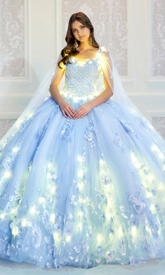 A young woman wears the PR22021 Princesa Glow-in-the-Dark Quinceañera Dress, featuring light blue fabric, floral appliqués, and integrated lights, as she stands before a white-patterned wall.