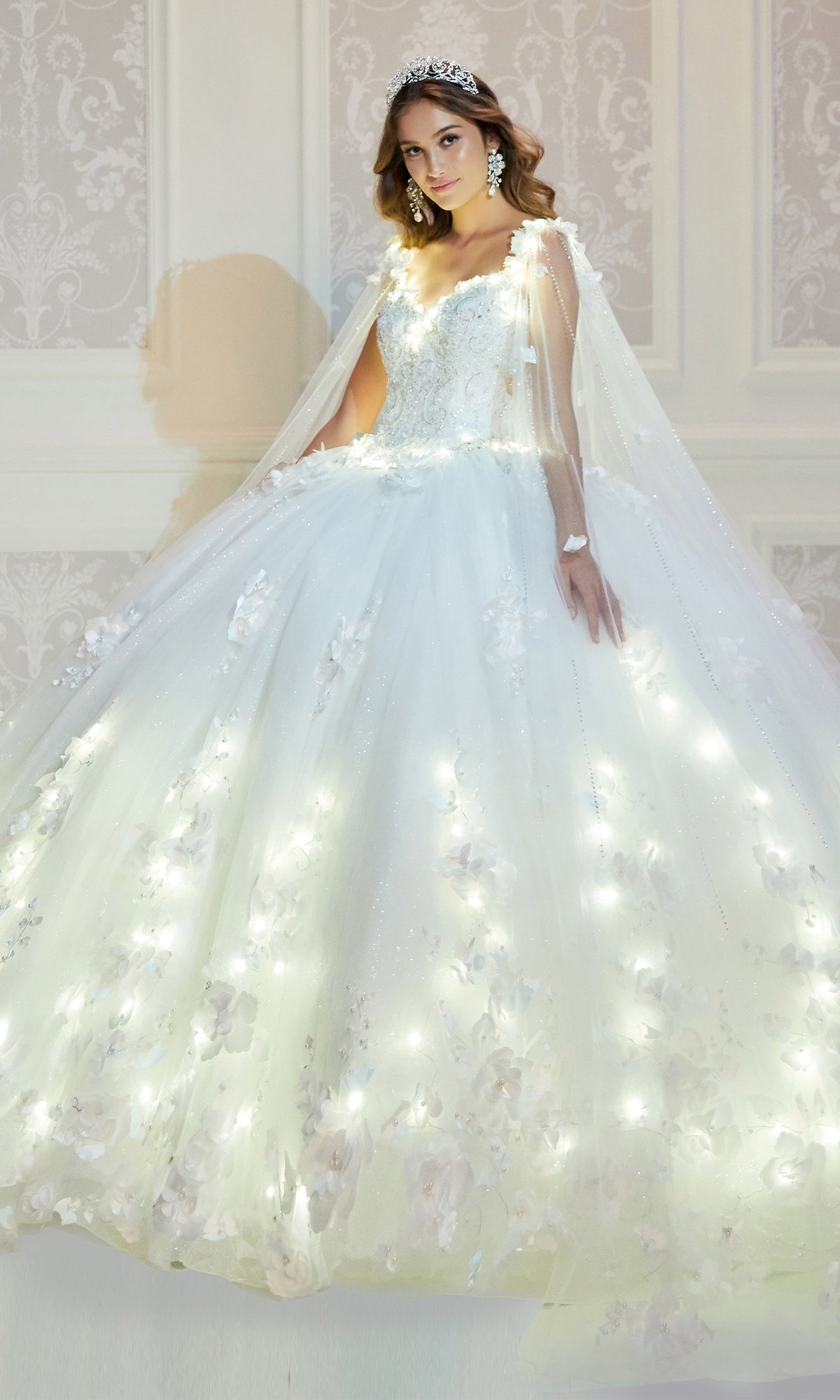 A woman stands indoors against an ornate wall wearing the PR22021 Princesa Glow-in-the-Dark Quinceañera Dress with floral embellishments and built-in lights, styled with a tiara and statement earrings.