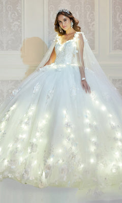 A woman stands indoors against an ornate wall wearing the PR22021 Princesa Glow-in-the-Dark Quinceañera Dress with floral embellishments and built-in lights, styled with a tiara and statement earrings.