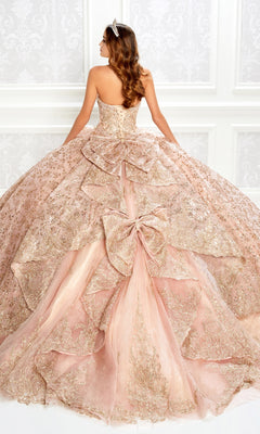 A woman in the Strapless Quinceanera Dress: Princesa PR22022—blush pink with gold details, large bows, and a tiara—stands facing away in an ornate room.