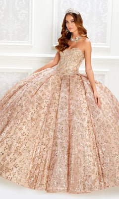 A woman stands elegantly against a white ornate background, wearing the Strapless Quinceanera Dress: Princesa PR22022 with gold sequins and a full skirt, paired with a tiara and statement necklace.