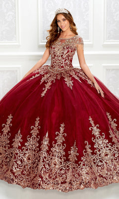 A woman wearing the Princesa PR22026 short-sleeve quinceanera dress with gold embroidery, a beaded bodice, and glitter tulle stands by a white ornate wall, smiling and accessorized with a matching tiara.