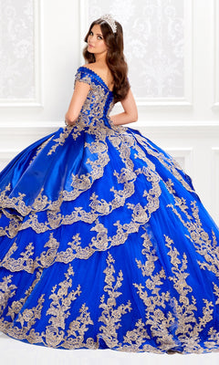 A woman in the Satin Quinceanera Dress: Princesa PR22029—a royal blue off-the-shoulder gown with gold embroidery—stands in an ornate white room, gazing over her shoulder.